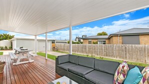 A covered patio area with a wooden deck, a grey sofa with cushions, a picnic table, and a small table with a plant. - Fielding's Lair - Pet Friendly (Port Fairy)