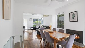 A modern living room with a dining area, a fireplace, and large windows offering a view of greenery. - Fielding's Lair - Pet Friendly (Port Fairy)