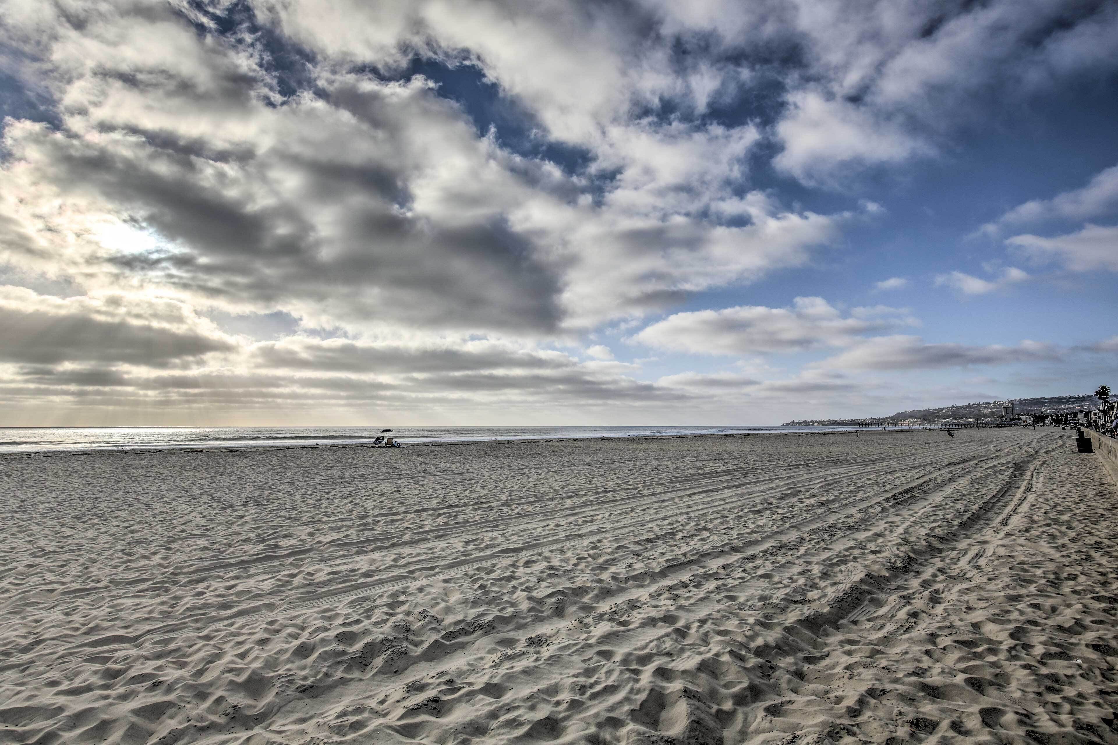 A wide sandy beach with footprints leading to the water, a pier in the distance, and a partly cloudy sky.