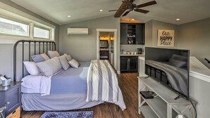 Ein Schlafzimmer mit Bett, Fernseher, Deckenventilator und einem durchgegangene Tür erkennbaren Küchenbereich.