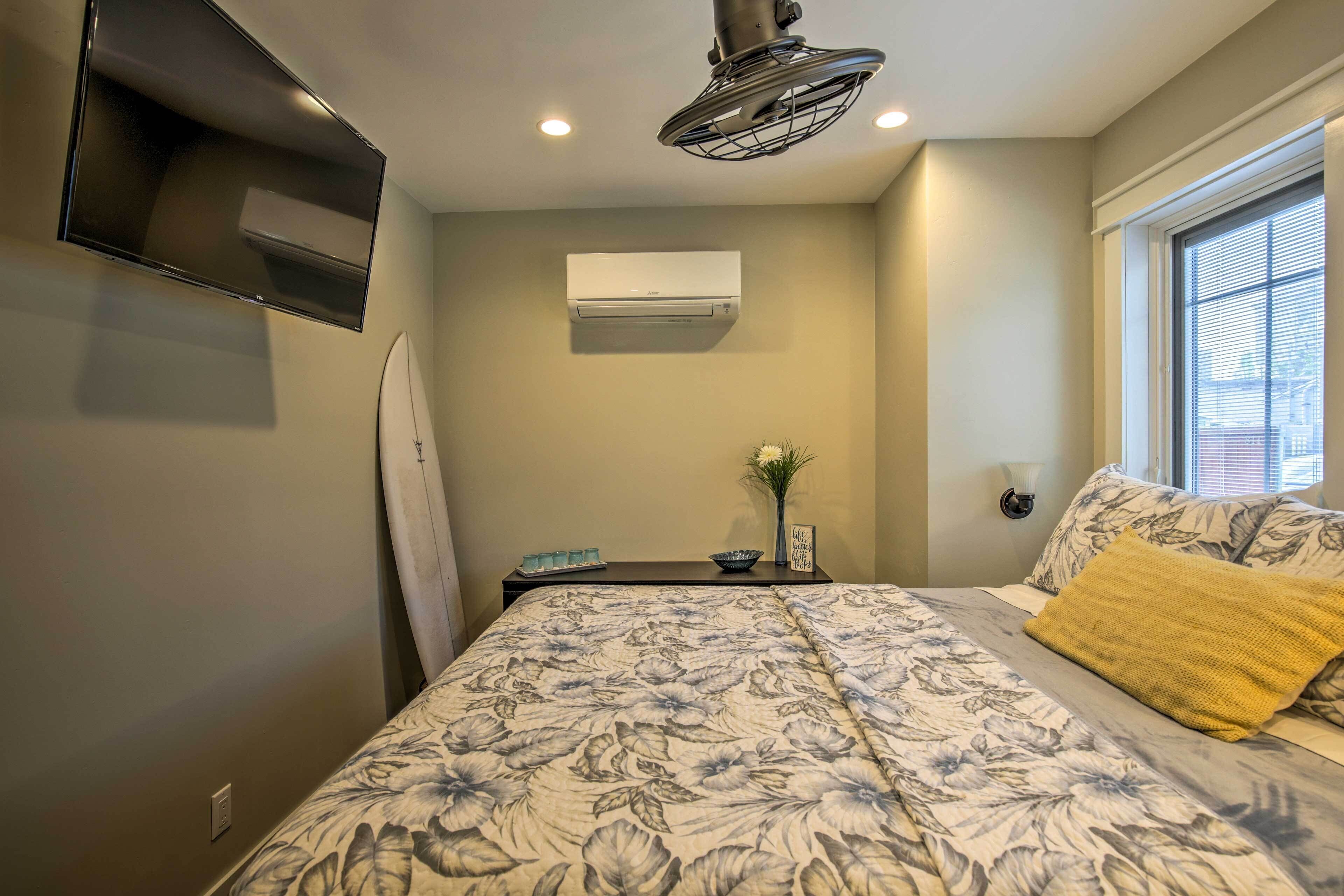 Una camera da letto con una coperta a fiori, una TV a parete, un condizionatore d'aria, un comodino con un vaso e un cartello, e una finestra con persiane.