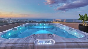 Un jacuzzi con panel de control, rodeado por una terraza de madera, con vistas a un paisaje urbano y al mar.