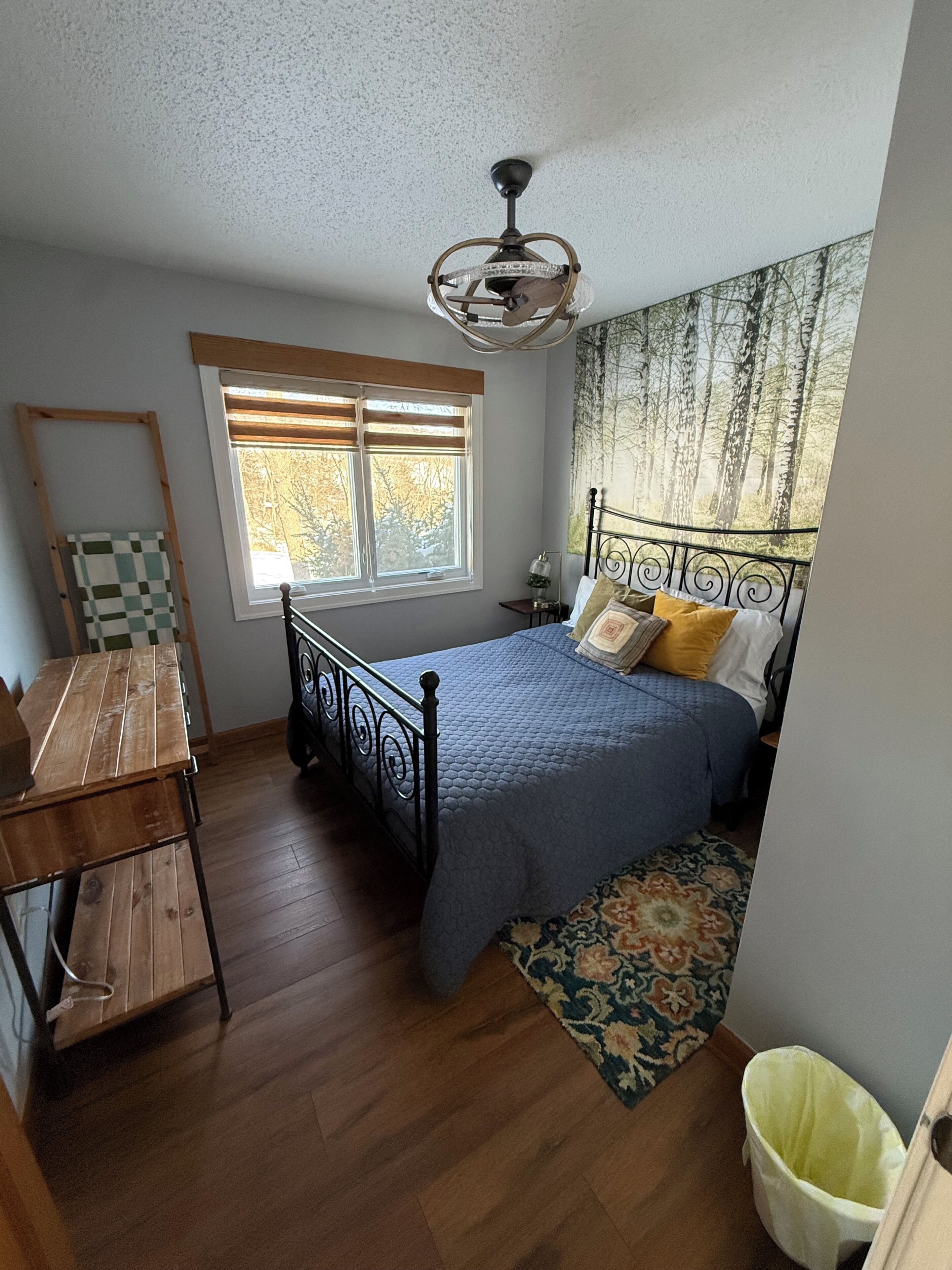 Une chambre à coucher comprenant un lit, un bureau, une fenêtre avec des stores, un ventilateur de plafond et une vue sur les arbres par la fenêtre.