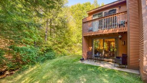 Una cabaña de madera de dos pisos con un balcón, un patio con mobiliario exterior y un bosque al fondo.