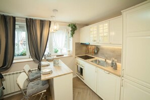 Une cuisine moderne avec des meubles blancs, un four encastré et un évier. On y trouve un coin repas avec une table et des chaises, ainsi qu’une fenêtre avec des rideaux.