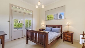 A bedroom with a wooden bed, two bedside tables with lamps, a mirror above the bed, a door with a view of greenery, and a small desk with a chair. - Situated in the CBD district with easy access to all Warrnambool has to offer (Warrnambool)