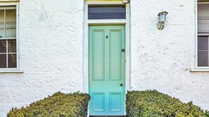 Un bâtiment blanc avec une porte verte, une fenêtre et un petit arbuste devant.