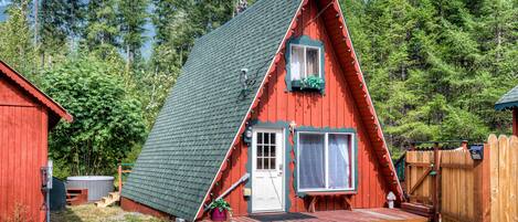A red A-frame cabin with a wooden deck, a white door, and a window with a curtain.