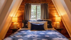 Un dormitorio con techo inclinado, paredes de madera, una cama grande con ropa de cama floral, dos lámparas de noche y una ventana con cortinas.