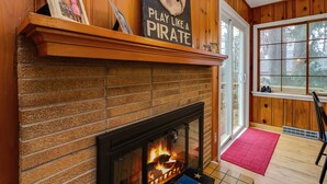 A fireplace with a brick surround, a mantel with a framed photo and a sign that says "Play Like a Pirate," and a window with a view of trees. - B and B's Beach House Accommodates Eleven Guests In 5 Bedrooms 2 Full Baths! (South Haven)
