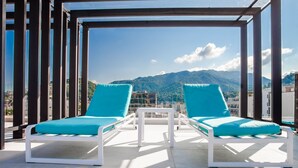 Eine Dachterrasse mit zwei weißen Liegestühlen und einem kleinen Tisch, mit Blick auf eine gebirgige Landschaft.