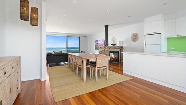 A modern kitchen with a stone fireplace, wooden dining table, and a view of the ocean. - Offering breathtaking views of South Beach (Port Fairy)