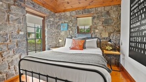 A bedroom with a stone wall, a bed with a metal frame, a nightstand, a lamp, and a window. - A bluestone cottage in the heart of Port Fairy (Port Fairy)