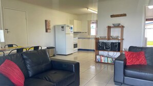 A living room with two black leather sofas, one with a red pillow. There is a kitchen area with a refrigerator, microwave, and a shelf with speakers and books.