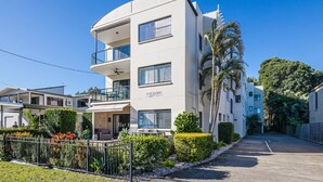 Un immeuble résidentiel moderne à plusieurs étages, doté de balcons, d’un espace extérieur couvert et d’un jardin bien entretenu.