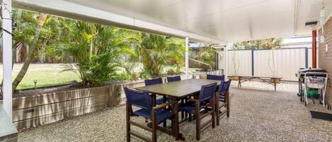 An outdoor patio with a wooden table and chairs, surrounded by gravel, with a covered area overhead and a view of a garden with palm trees.