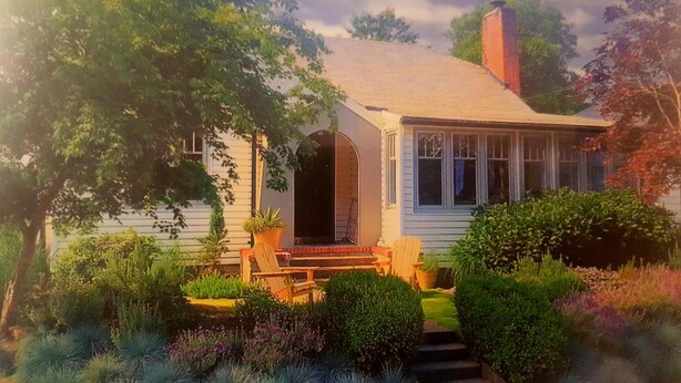 Einbungalow mit einer Veranda, einem Schornstein und einem Garten mit verschiedenen Pflanzen und Sträuchern.
