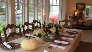 Ein Esszimmer mit einem langen Tisch, gedeckt für eine Mahlzeit, mit hölzernen Stühlen, einer weißen Tischdecke und einer Mittelstück-Dekoration aus Blumen und Kürbissen.