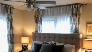 Ein Schlafzimmer mit einem großen Bett, zwei Nachttischlampen, einem Deckenventilator und Vorhängen.