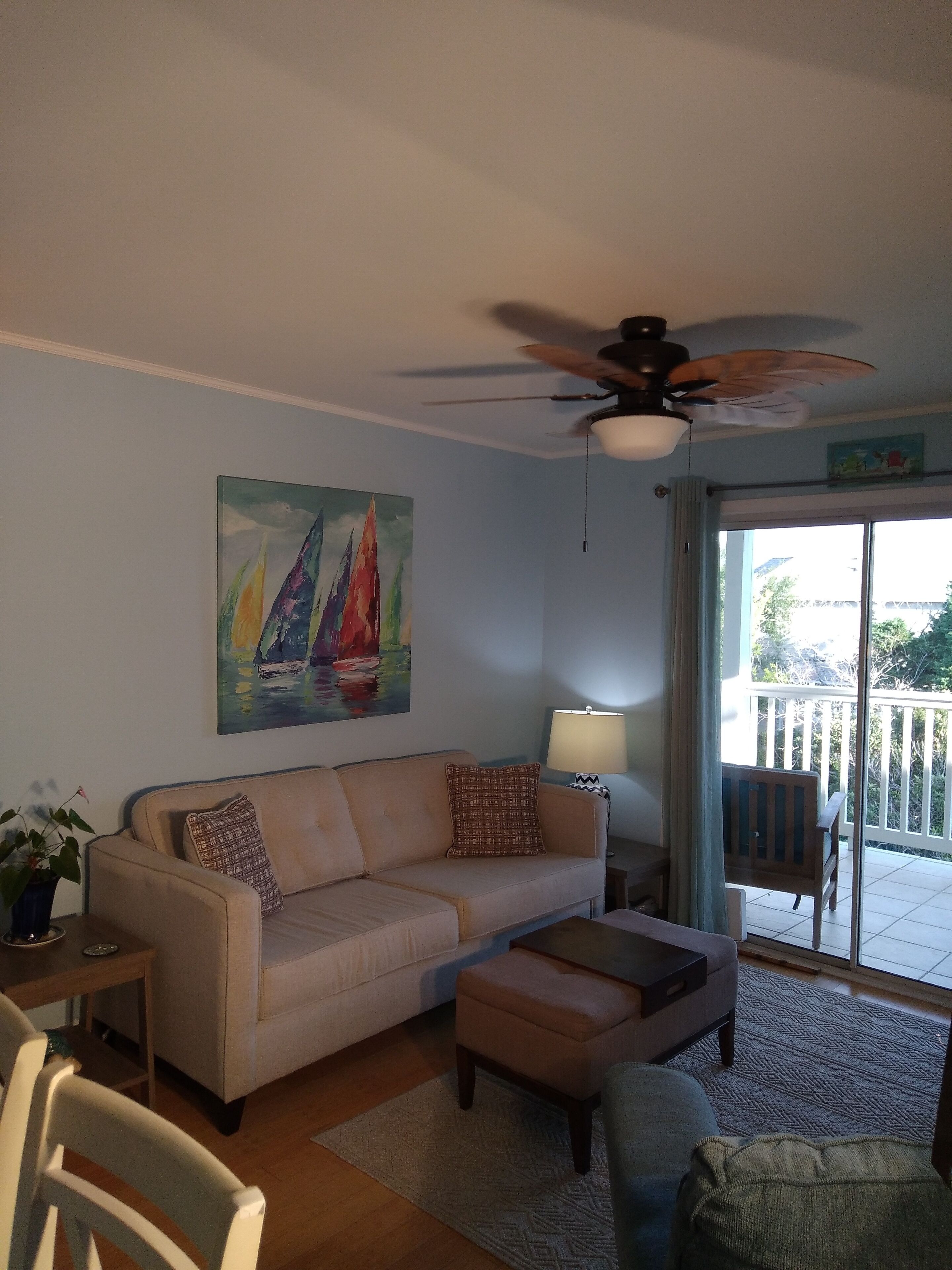 Un salon comprenant un canapé, une table basse, un ventilateur de plafond, un tableau et une vue sur l’extérieur grâce à une porte vitrée.