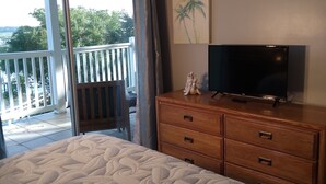 A bedroom with a large bed, a wooden dresser, a television, and a view of a balcony with a railing.