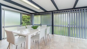 Un comedor moderno con una mesa y sillas blancas, un cuenco verde y vistas amplias a través de ventanas con persianas.
