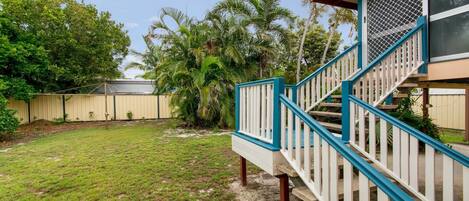 A residential backyard with a staircase leading to a covered porch, surrounded by palm trees and a fence.