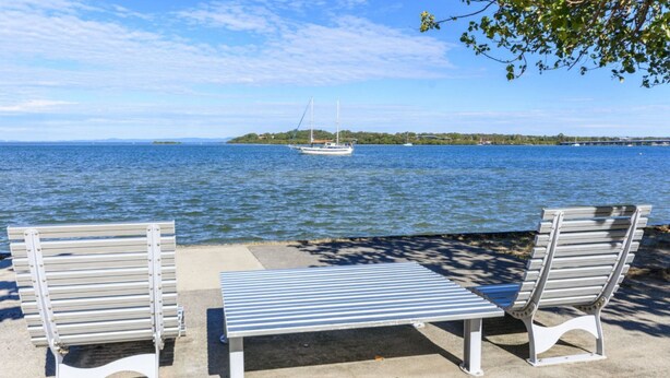 Two metal benches and a table on a concrete surface by a waterfront with a sailboat in the distance. - Charm and Comfort on Bribie (BONGAREE)