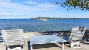 Two metal benches and a table on a concrete surface by a waterfront with a sailboat in the distance.