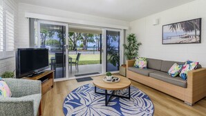Een moderne woonkamer met een flatscreen-tv, een grijze bank met kleurrijke kussens, een ronde salontafel en een grote schuifpui die uitkomt op een balkon met uitzicht op zee en groen.