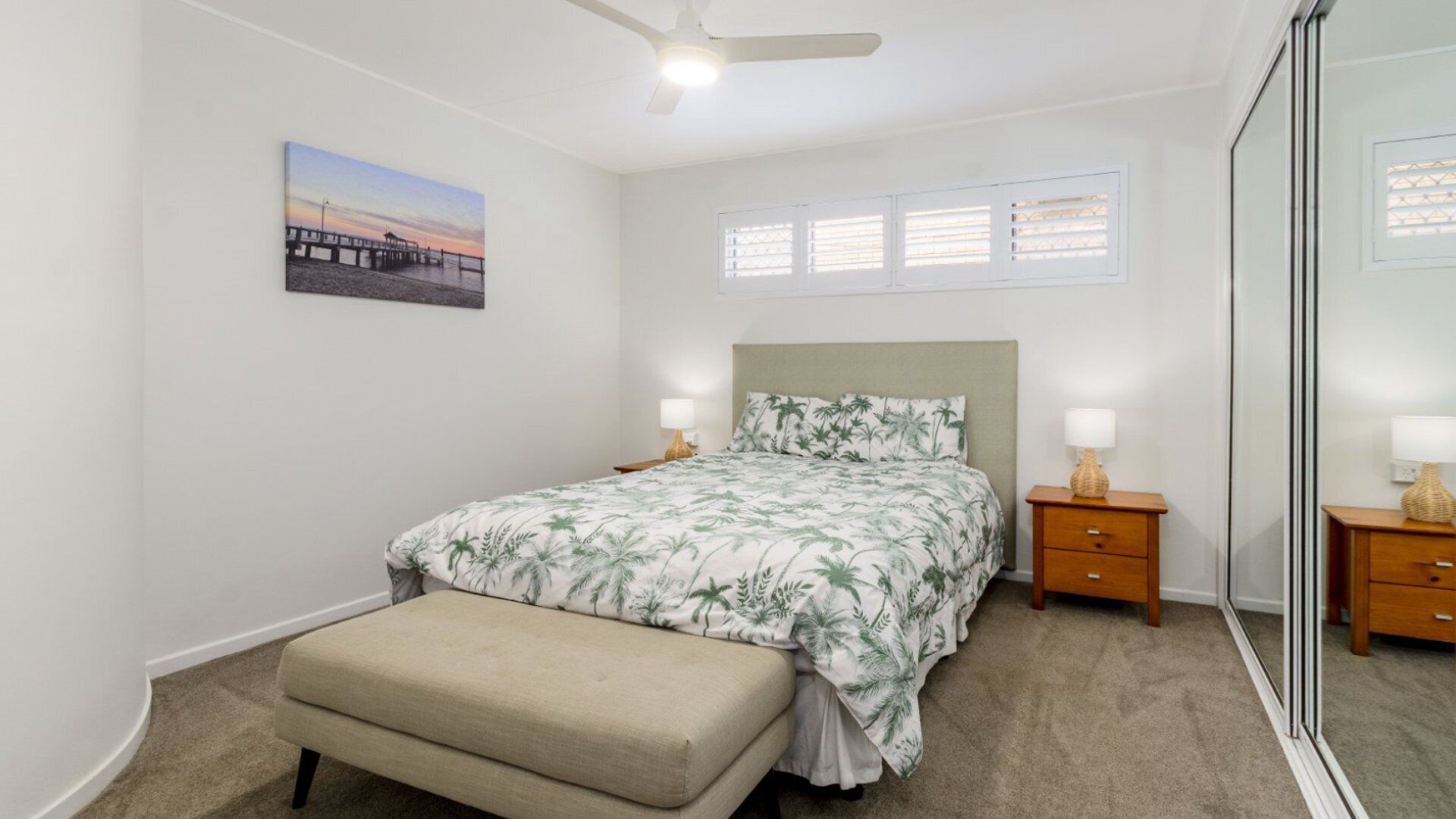 Une chambre à coucher comprenant un lit, des tables de chevet, un banc, un ventilateur de plafond, une fenêtre avec des stores et un tableau accroché au mur.