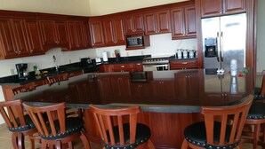 Una cocina con armarios de madera, un refrigerador de acero inoxidable, un microondas, una cafetera y una estufa. Hay seis taburetes de bar de madera con asientos negros.