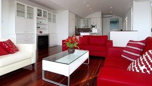 Una sala moderna con un sofá rojo, un sofá blanco y una mesa de centro de vidrio. Se observa una cocina al fondo con armarios blancos y un microondas.