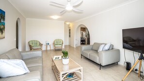 A living room with two sofas, a coffee table, and a TV. There are two armchairs and a small table with decorative items. The room has a ceiling fan and a view of a dining area through an archway.