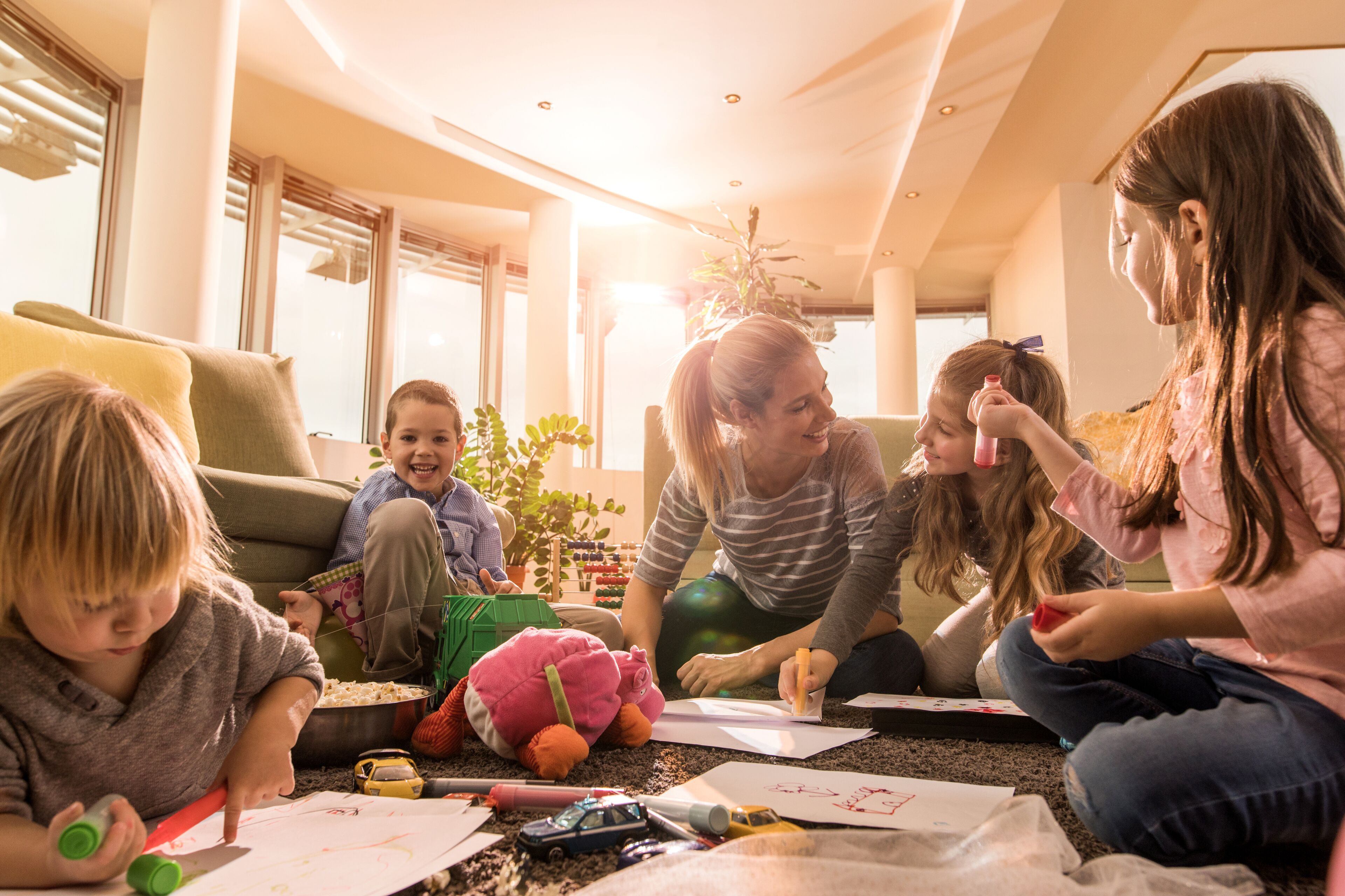A group of children and an adult sitting on the floor in a well-lit room with large windows and a modern interior.