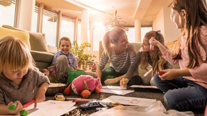Un groupe d’enfants et un adulte sont assis sur le sol dans une pièce bien éclairée, dotée de grandes fenêtres et d’un intérieur moderne.