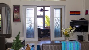 Un salon avec un téléviseur, une table basse avec des livres et une petite plante, un coin repas avec des chaises et une table, et un balcon avec vue.