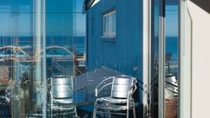 Un balcone con un tavolo e sedie in vetro, affacciato su un edificio blu e sulla vista del mare.