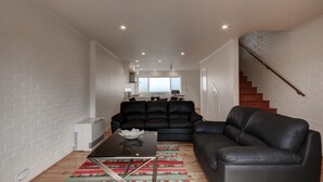 Ein modernes Wohnzimmer mit einer schwarzen Ledersofa, einem Glastisch und einem gemusterten Teppich. Eine Treppe befindet sich rechts und im Hintergrund eine Küche.