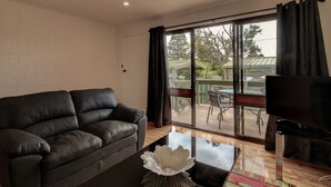 Ein Wohnzimmer mit schwarzem Ledersofa, einem Glastisch mit dekorativer Schale, einem Flachbildfernseher und einer Schiebetür zu einem Außenbereich mit Tisch und Stühlen.