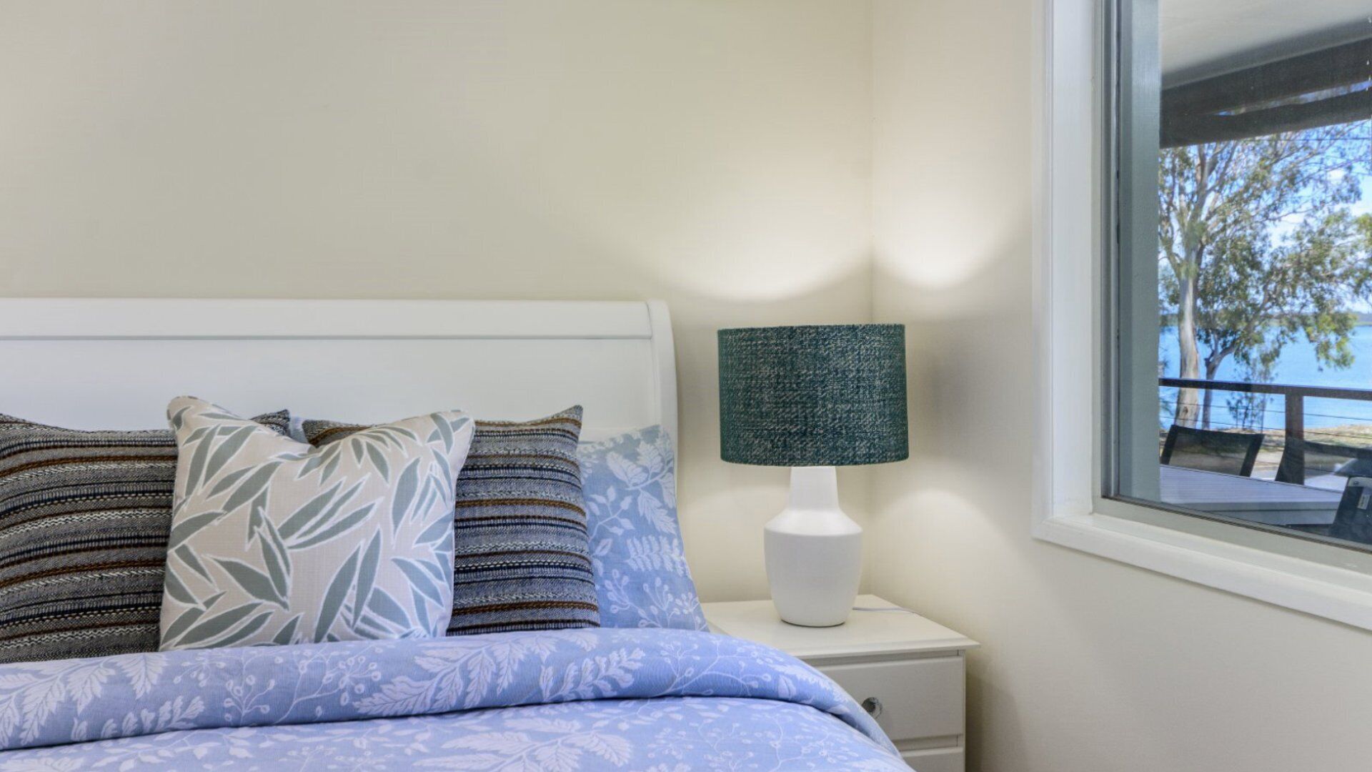 A bedroom with a bed, pillows, a nightstand, a lamp, and a window with a view of trees and a balcony.