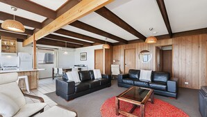 A spacious living area with a black leather sofa, a wooden coffee table, and a red carpet. There is a kitchen area with white appliances and a dining table. The room features exposed wooden beams and a large window. - Perfect for that cosy winter holiday with stunning views of the Pea Soup Cove. (PORT FAIRY)