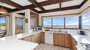 A kitchen with wooden cabinets, a white countertop, a microwave, a toaster, and a large window with a view of the beach. - Perfect for that cosy winter holiday with stunning views of the Pea Soup Cove. (PORT FAIRY)