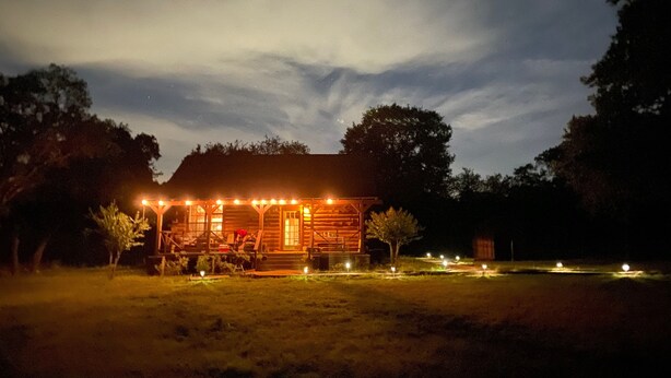 Un chalet rustique en rondins de bois, doté d’un porche illuminé par des guirlandes lumineuses, et entouré d’arbres.