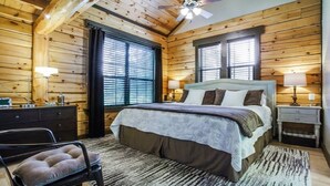 Une chambre confortable dans un chalet en rondins, avec un grand lit, deux tables de chevet avec des lampes, un fauteuil et un ventilateur de plafond.