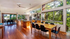 Un vaste espace dédié à la salle à manger, avec une longue table en bois et des chaises noires, de grandes fenêtres offrant une vue sur la verdure, et un coin cuisine équipé d’appareils électroménagers modernes.