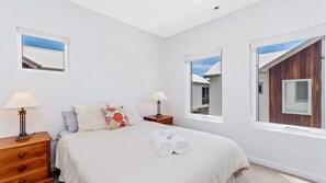 Une chambre à coucher comprenant un lit, deux tables de chevet avec des lampes, une commode et des fenêtres donnant sur les bâtiments voisins.