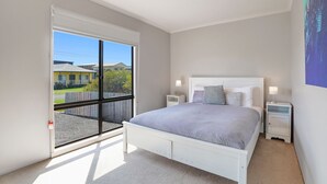 Une chambre avec une grande porte-fenêtre coulissante donnant sur un balcon, un lit blanc avec une tête de lit grise, deux tables de chevet avec des lampes et une vue sur les maisons à l’extérieur.