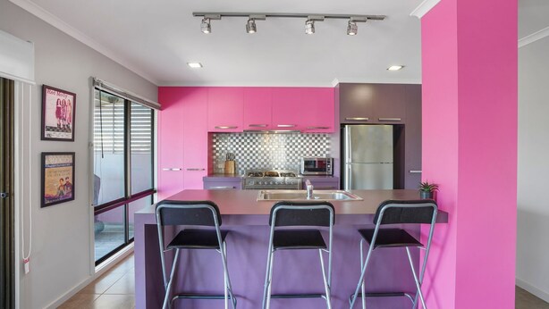 Une cuisine moderne avec des meubles de couleur rose, un réfrigérateur en acier inoxydable, un four à micro-ondes et une cuisinière à gaz. On y trouve trois tabourets de bar et une fenêtre avec des stores.