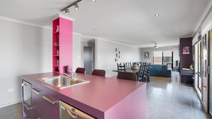 Une cuisine moderne avec un îlot central rose, un coin repas avec un canapé bleu et un salon doté d’une grande fenêtre.